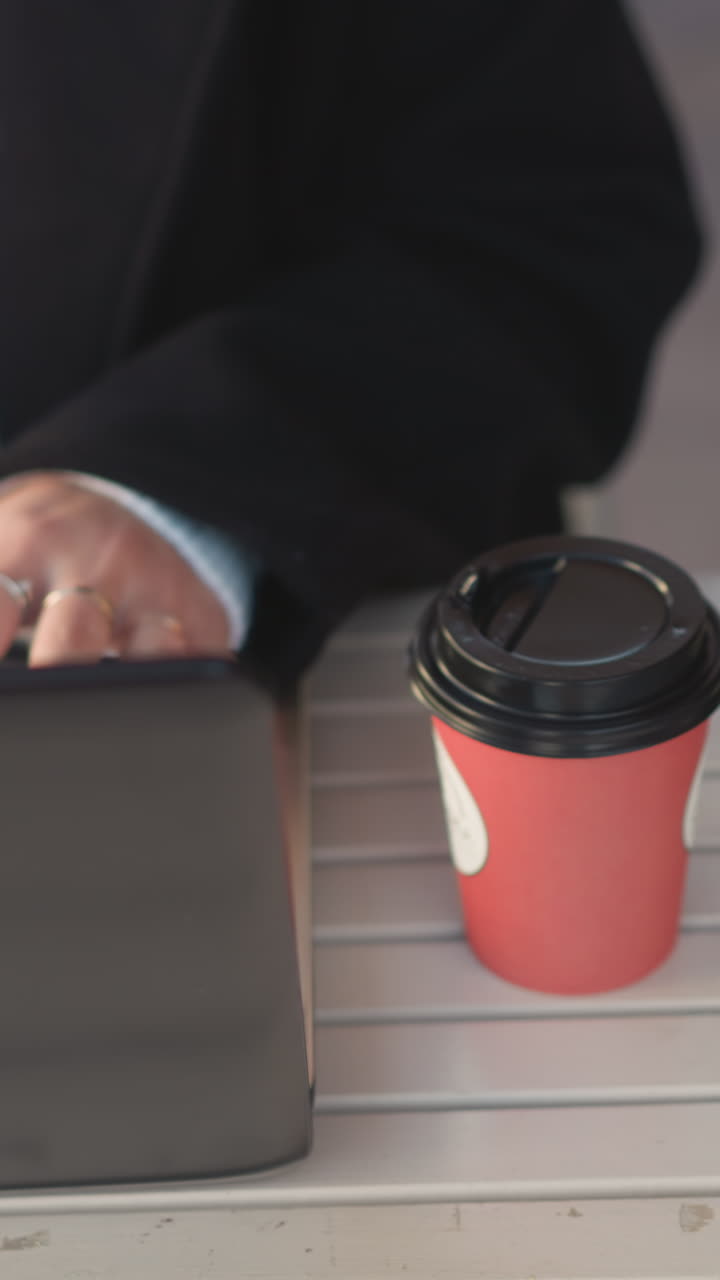 primer plano de las manos escribiendo en el teclado de la computadora portátil en una mesa al aire libre, persona con abrigo negro, suéter blanco y anillos mientras la taza de café está al lado de la laptop