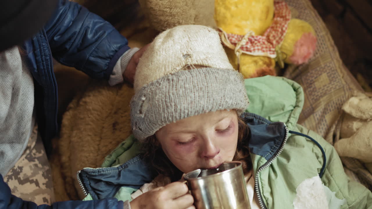 Top down view of weak child wearing torn hat and jacket slowly drinking water from metal cup while resting, showing physical exhaustion, cold environment, and emotional moment of care and survival