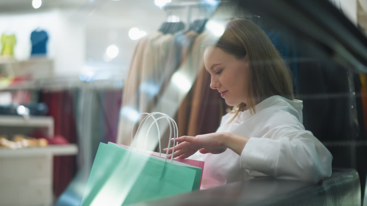 소파에 앉아있는 여성의 클로즈업, 다양한 색상의 쇼핑 가방을 들고, 쇼핑몰에서 가방 안에 있는 물건을보고, 분홍색, 초록색, 파란색의 가방이 그녀의 무에 놓여있는 동안 그녀는 휴식을 취합니다.