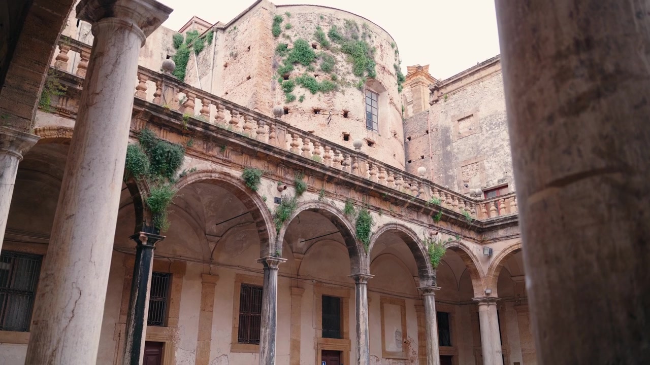 Un cortile storico in pietra incorniciato da archi a tutto sesto e colonne rivela una torre coperta d'edera che si erge sopra, in Sicilia.