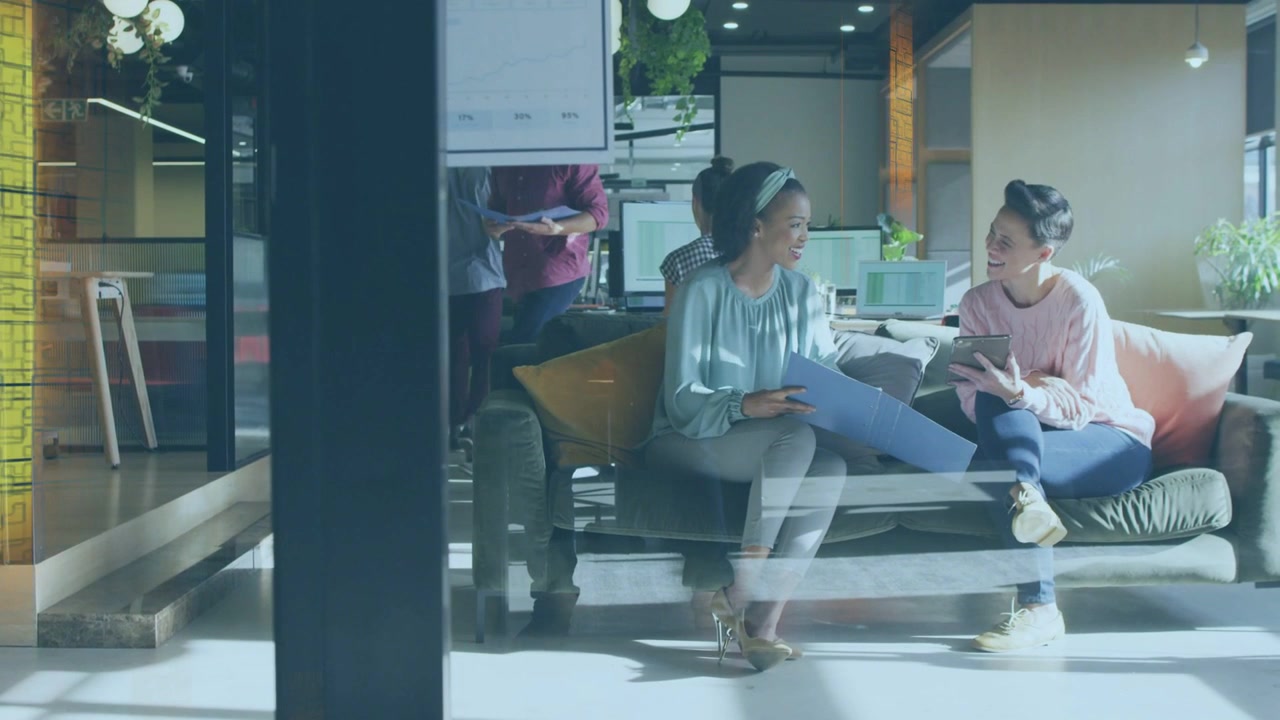 オフィスラウンジで、ピンクのセーターと青いブラウスを着た2人の女性がタブレットをタップしながらフォルダーを確認している。