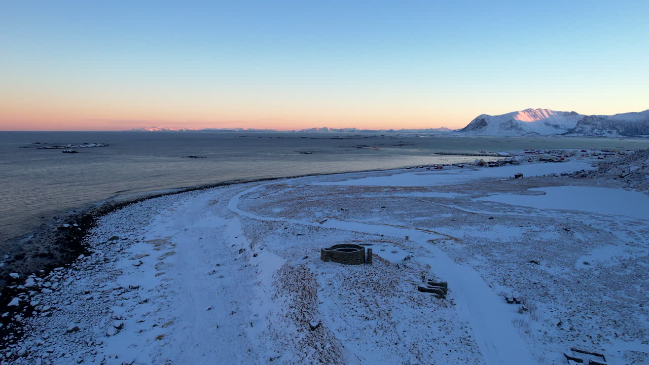 Cinematic 4K drone push in over the historic ruins in Eggum. Dramatic winter landscape with snowy coast and mountains during Arctic blue hour sunrise in Lofoten, Norway