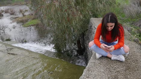 young woman sitting by a river, looking at her phone