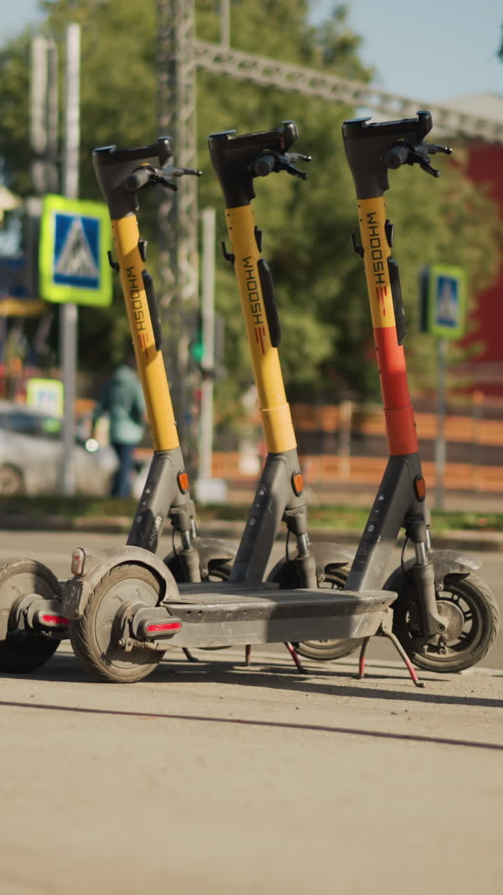 row parked electric scooters on city pavement near crosswalk sign, sunlit roadside with blurred background, rental branding and QR codes visible, kickstands down, modern mobility solution