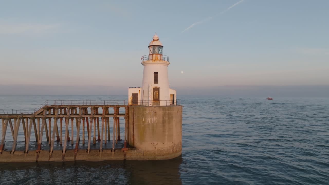 Scenic lighthouse, calm sea and jetty at golden hour, with a full moon and boat in background