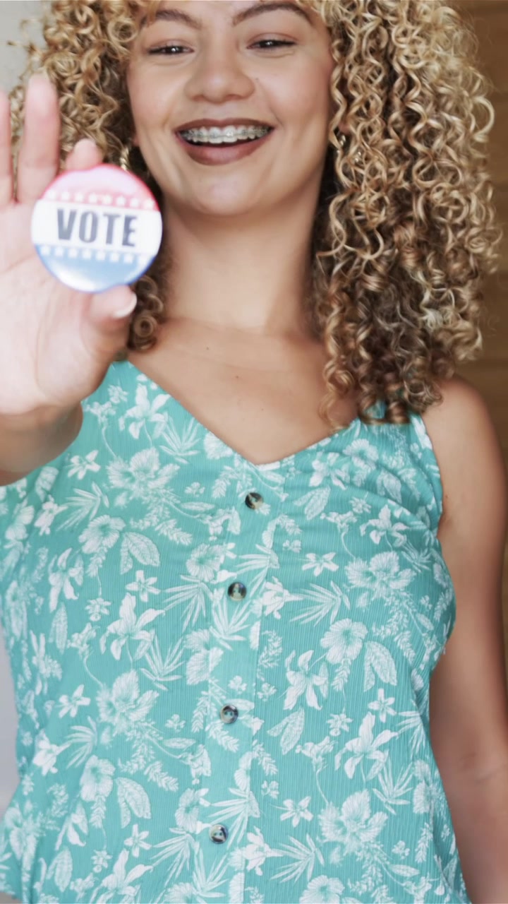 Video verticale: Donna sui vent'anni che cammina da sinistra nell'ingresso, mostrando una spilla 'VOTA' e incoraggiando il voto