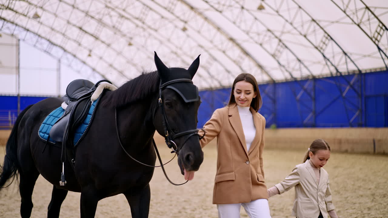 Beautiful long-haired lady in beige jacket leading a black horse in ammunition. Little girl walking by holding a woman by the hand. Manege at backdrop in blur.