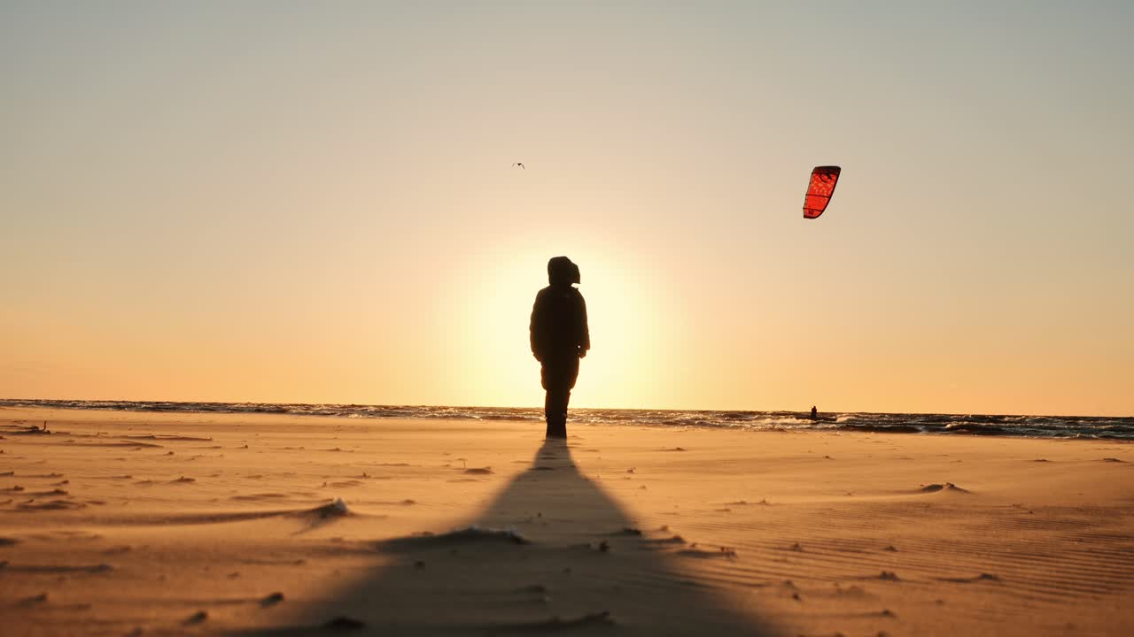 Lonely figure stands on Baltic coast as kite surfer sails under warm sunset sky