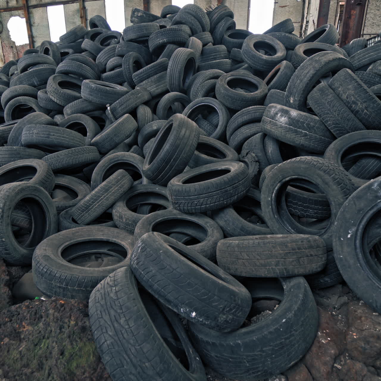 Big pile of automobile tires on the broken-down plant with holes in the wall and roof. Many black rubber tyres on the ground inside the old huge empty building. Motion camera bottom up.