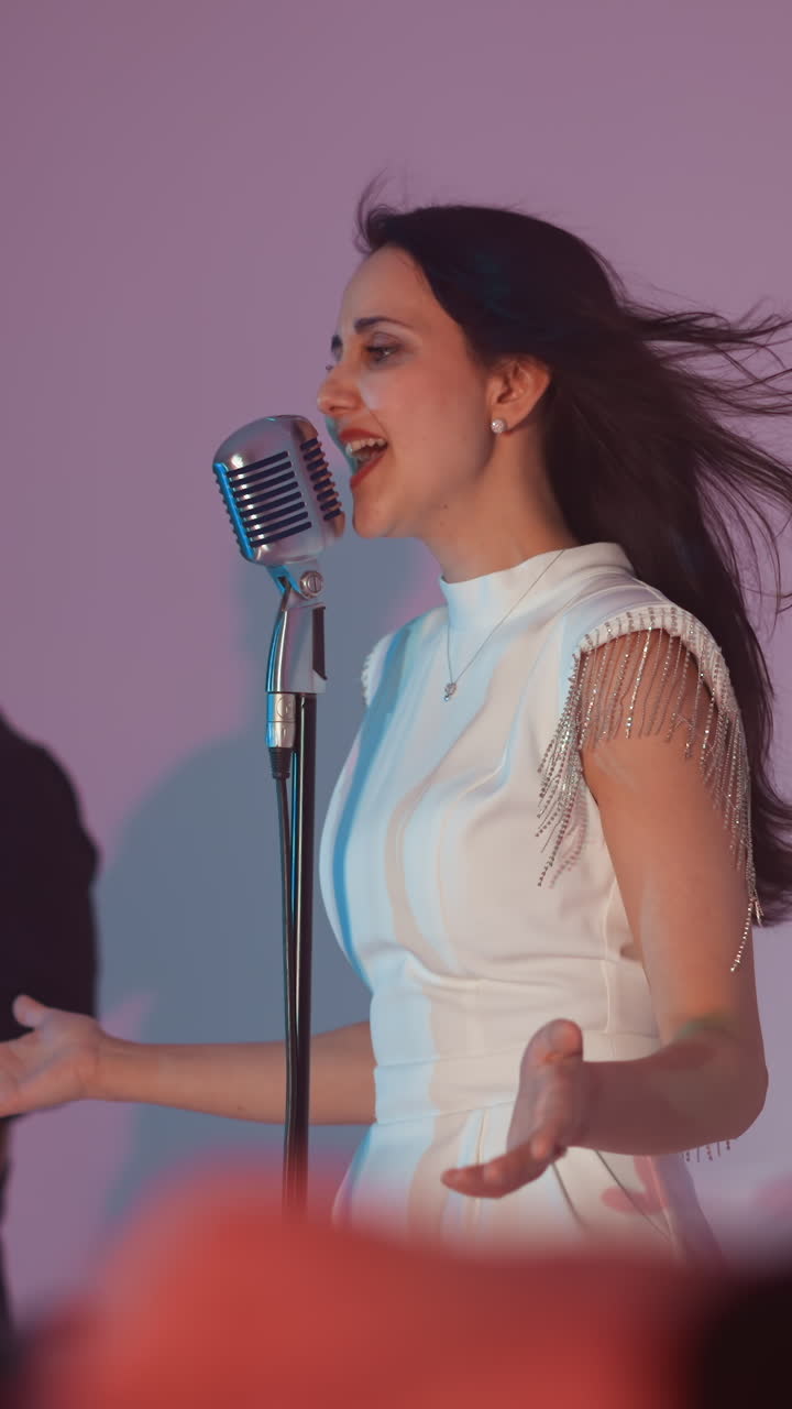 A female singer wearing a white gown stands in front of a vintage microphone, passionately singing. A guitarist plays in the background