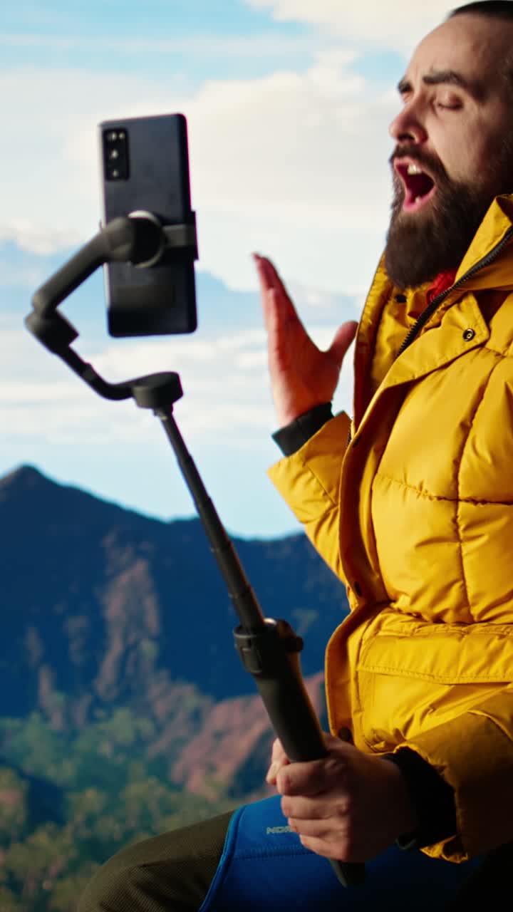 Uomo barbuto che fa vlogging con smartphone su selfie stick in montagna