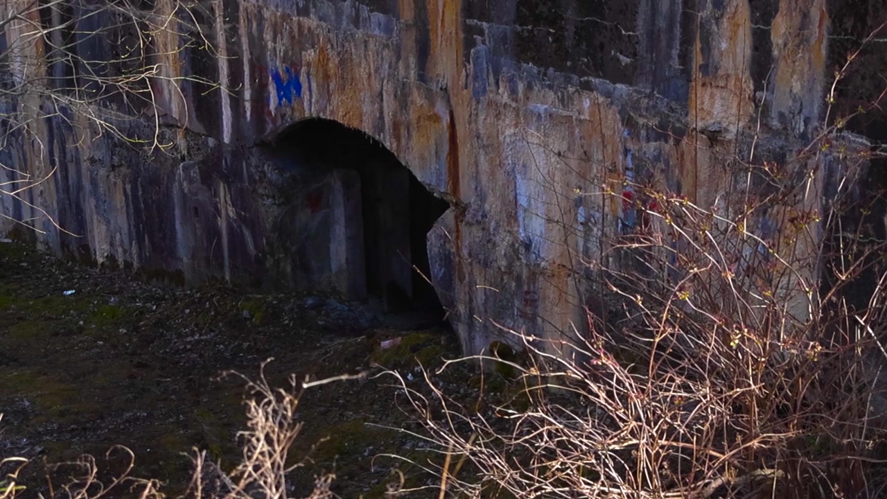 Antiga muralha de pedra da Fortaleza Naval de Pedro, o Grande, mostrando uma entrada de caverna escura em arco, com texturas desgastadas e arbustos secos banhados pela luz solar intensa em um dia tranquilo.