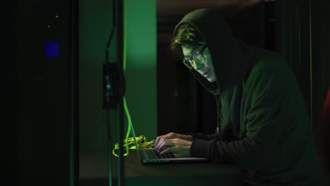 asian male hacker in hoodie using laptop by computer servers