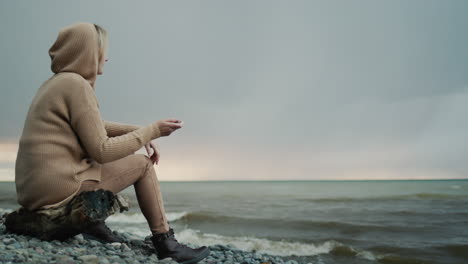 alone woman in a warm sweater with a hood on her head sits on the ocean shore, where the dramatic sky and surf