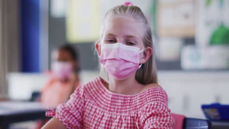 Portrait-of-caucasian-schoolgirl-wearing-face-mask,-sitting-in-classroom-looking-at-camera