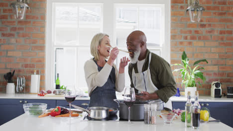 Pareja-De-Ancianos-De-Raza-Mixta-Usando-Delantales-Probando-Comida-Mientras-Cocina-En-La-Cocina-De-Casa