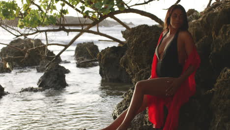 latino girl model poses stroking her legs skin sitting on tropical island rocks, looks at camera