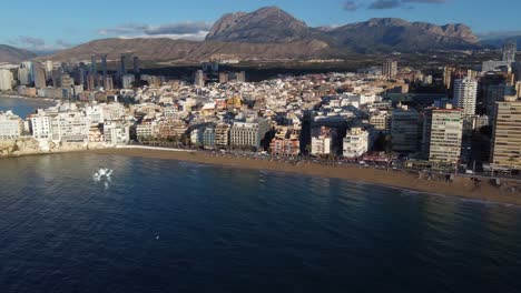 Toma-Panorámica-Aérea-De-La-Ciudad-De-Benidorm-Con-Torres,-Playa-De-Arena-Y-Cordillera-En-El-Fondo-Durante-El-Amanecer-Dorado