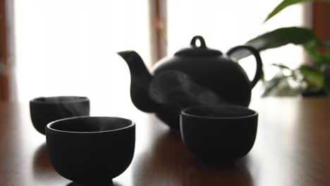 minimal background of a green japanese tea set with steam coming out of the cups, on a wooden table, with a plant and a window in the back