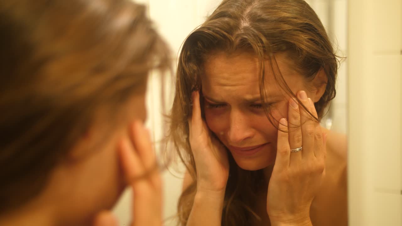 una mujer con moretones y cicatrices en la cara por haber sido golpeada y golpeada se mira en el espejo del baño y se limpia las lágrimas de la cara