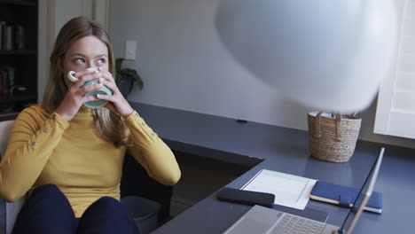 mujer biracial sentada en un escritorio usando una computadora portátil y bebiendo café en casa, en cámara lenta