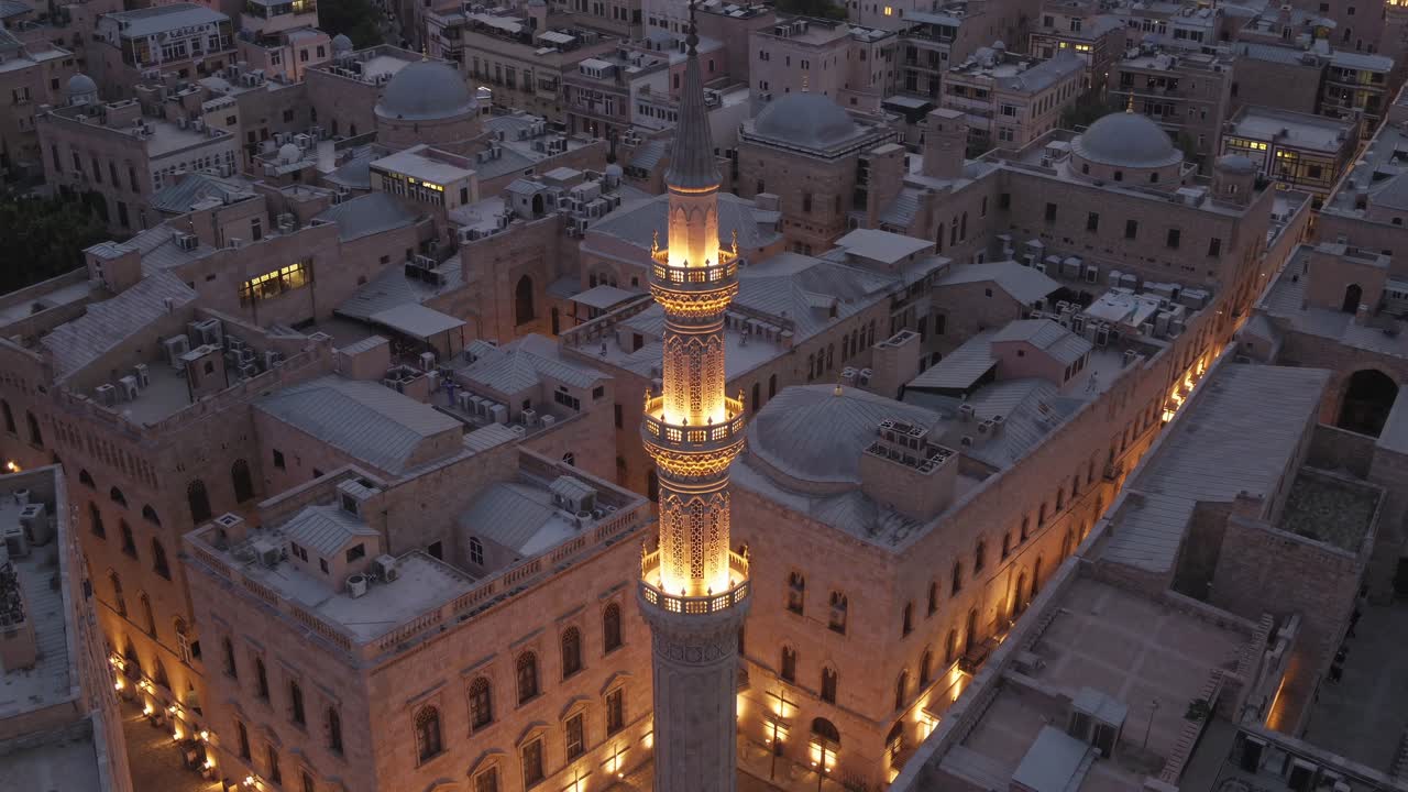 Illuminated minaret rises above a dense cityscape of traditional stone buildings at dusk, creating a serene atmosphere reminiscent of Ramadan, with warm lights adding to the tranquil ambiance