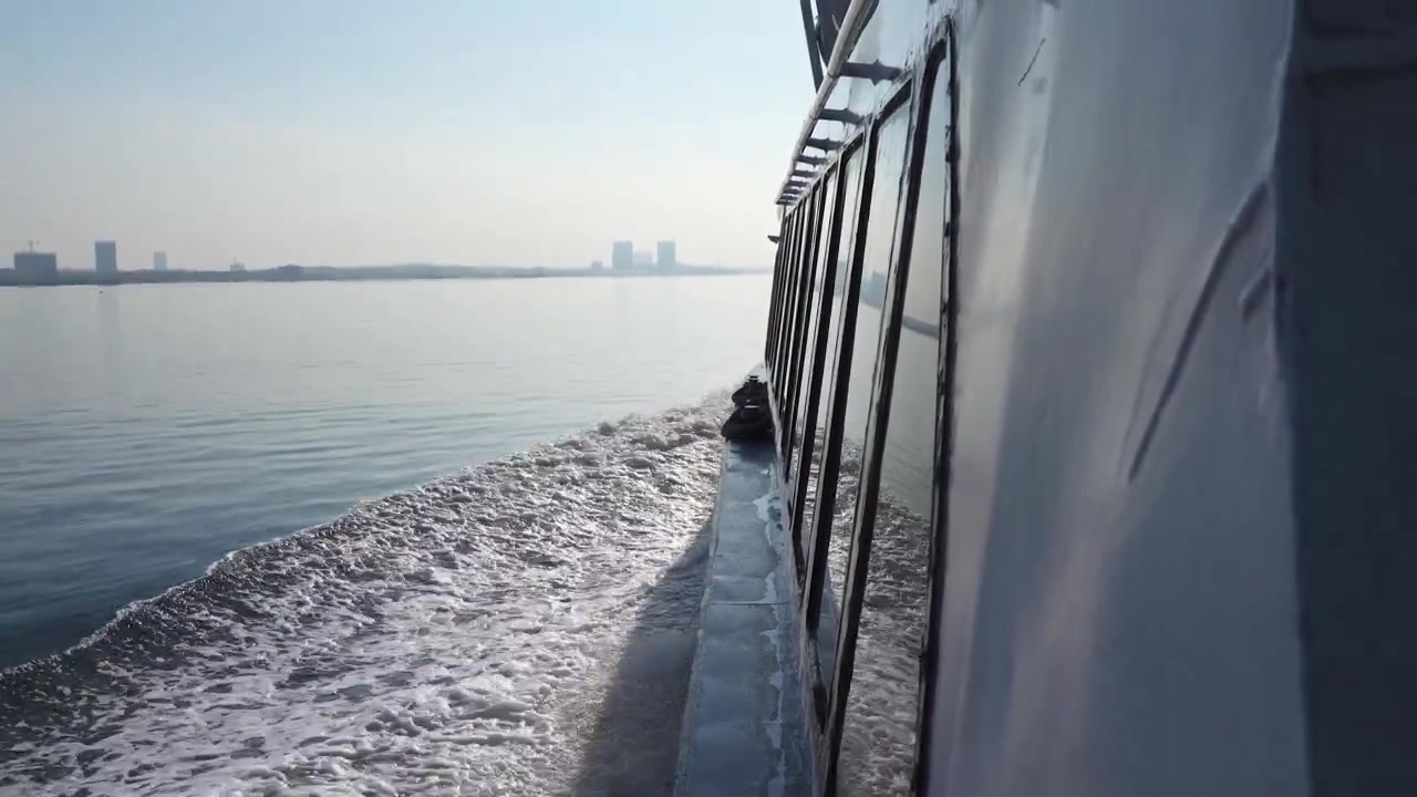 Eine Aufnahme aus der Ich-Perspektive vom Deck einer fahrenden Fähre, die eine weiße Schaumspur und eine entfernte Stadtsilhouette entlang einer wichtigen Schifffahrtsroute in Südostasien oder der Straße von Malakka zeigt. Reise- und maritime Themen.