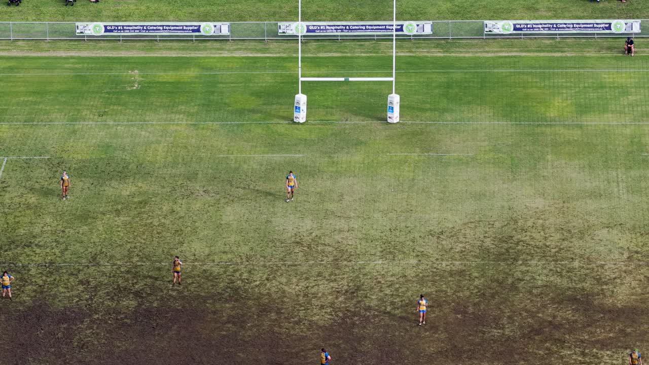 Children engage in a rugby match on a muddy field under bright daylight, captured from an aerial perspective in Gold Coast, Australia