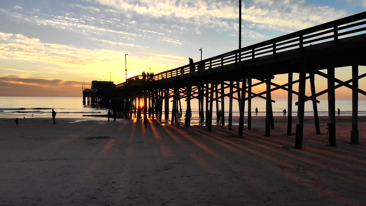 gente en silueta caminando por debajo y en el muelle de newport beach en el océano con haces de luz naranja al atardecer en la costa sur de california