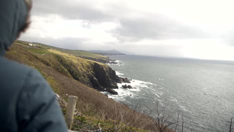 person in green hooded jacket stands on edge and watches towards cliffs and wild sea in distance, waves crash on shore, tourist in dingle peninsula, ireland, on cloudy, windy day, slow motion