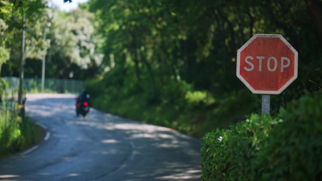 平和な緑の道の脇にある止まれの標識のクローズアップ。遠くをバイクが通り過ぎ、柔らかい自然な夏の光が差し込んでいる。