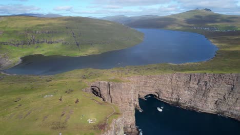 Hikers-trek-across-Lake-Sorvagsvatn-and-Traelanipa-edge-of-sheer-ocean-cliffs,-aerial-establishing