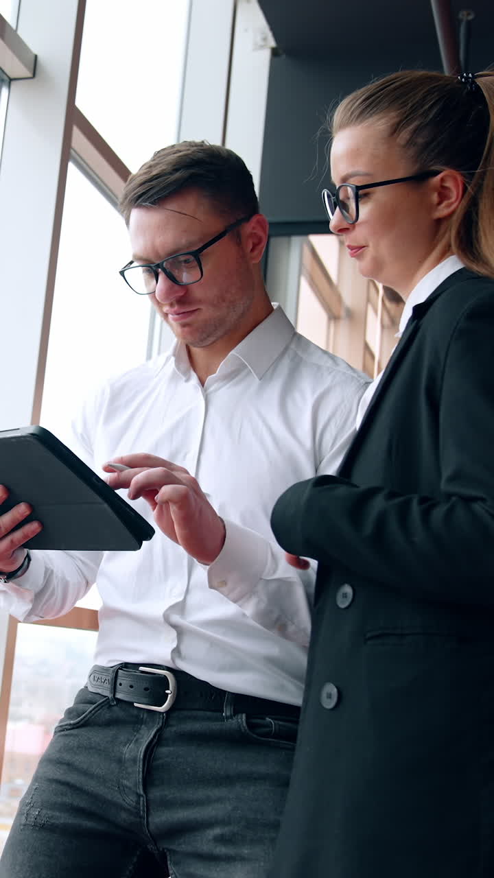 Female and male employees are in the office. Woman looks at I-pad in hands of man working on it. Low angle view. Vertical video