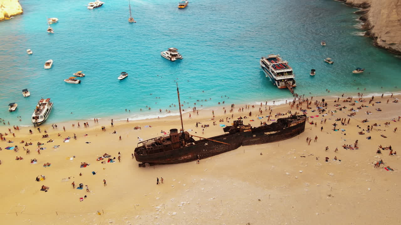 Aerial drone view of the Navagio beach on the Ionian Sea coast of Zakynthos, Greece. Moored boats and resting people, rusty ship, blue water. Slow motion