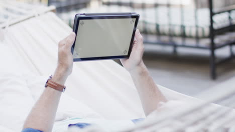 Kaukasischer-Mann-Mittleren-Alters,-Der-Ein-Tablet-Benutzt-Und-In-Der-Hängematte-Auf-Der-Terrasse-In-Der-Natur-Entspannt,-Zeitlupe