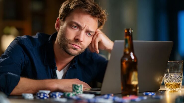 A contemplative man, engrossed in an online game, stares intently at his laptop screen amidst scattered poker chips and a refreshing drink, embodying the challenges and excitement of digital gambling
