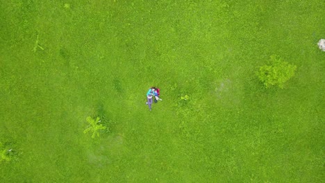 curly-hair-girl-with-a-young-boy-lie-down-on-green-field-with-fresh-grass-and-smile-happy-face-in-a-drone-shot-birdseye-shot-fly-over-take-off-the-ground-to-the-sky-wide-landscape-view