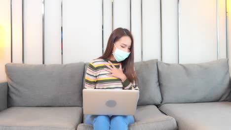 Asian-woman-have-a-heavy-cough-while-being-on-self-quarantine-during-covid-19-virus-pandemic,-she-is-using-her-laptop-computer-sitting-on-the-grey-couch-at-home