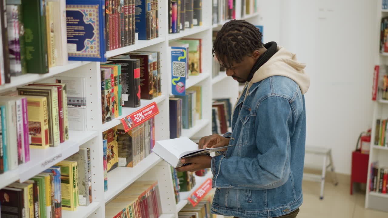 Young man browsing bookshelves in modern library, holding book, focusing on selection, surrounded by colorful book covers, bright indoor lighting, cozy library atmosphere