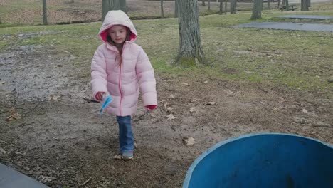 a young girl walks from behind a tree in the park during a light rain to pick up trash and throw it in a blue trash can