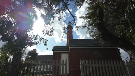 En-Medio-Del-Tranquilo-Entorno-Al-Aire-Libre,-Se-Alza-Un-Majestuoso-Edificio-De-Ladrillo-Con-Un-Faro,-Enmarcado-Por-Una-Valla-Blanca-Y-Adornado-Con-Ventanas-Que-Ofrecen-Vislumbres-Del-Vasto-Cielo-Y-Las-Nubes-Pasajeras.