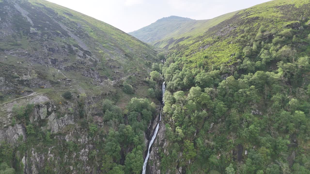 Zoom out drone footage reveals waterfall cascading down majestic mountain during daytime in England, UK. As camera pulls back, full scale of rocky terrain and surrounding landscape becomes visible