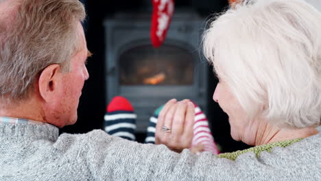 Rückansicht-Eines-älteren-Paares,-Das-Zu-Hause-Auf-Dem-Sofa-Am-Kaminofen-Sitzt-Und-Sich-Mit-Weihnachtsdekorationen-Im-Hintergrund-Unterhält