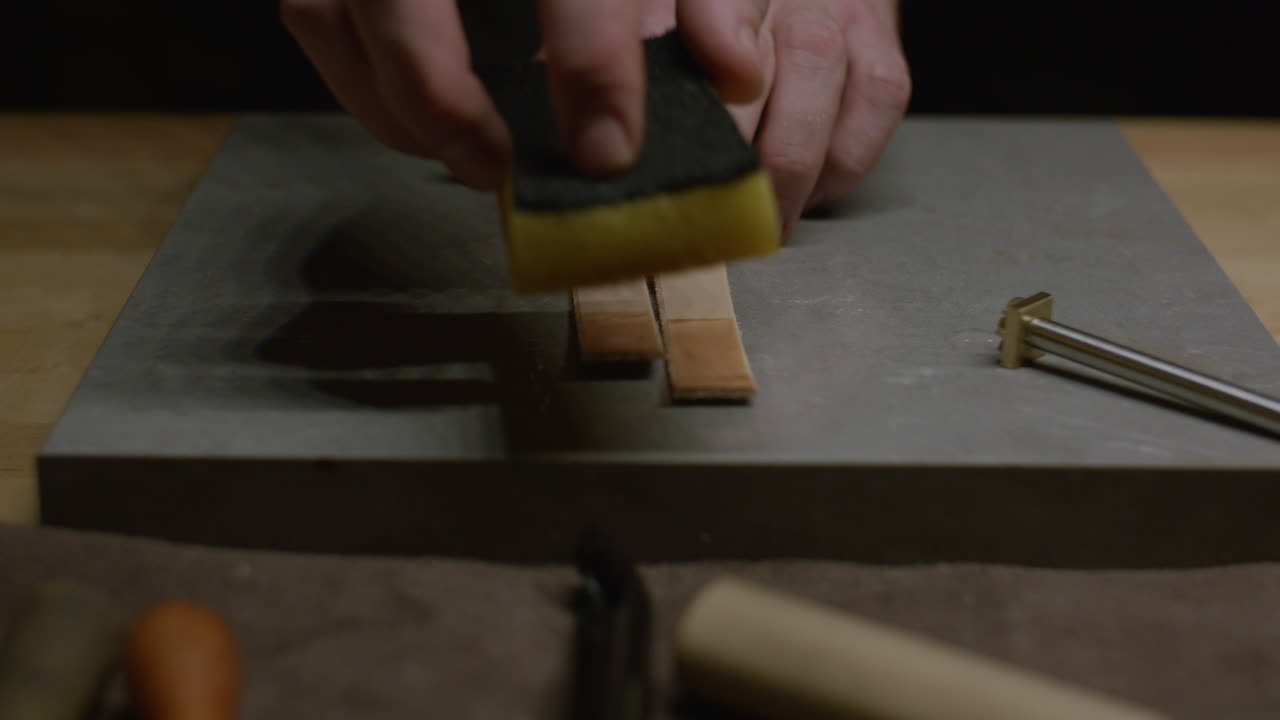 A leatherworker wets two small leather straps with a sponge, applying an even coat of water on a marble surface.