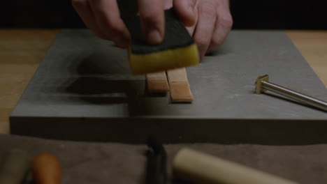 a leatherworker wets two small leather straps with a sponge, applying an even coat of water on a marble surface.