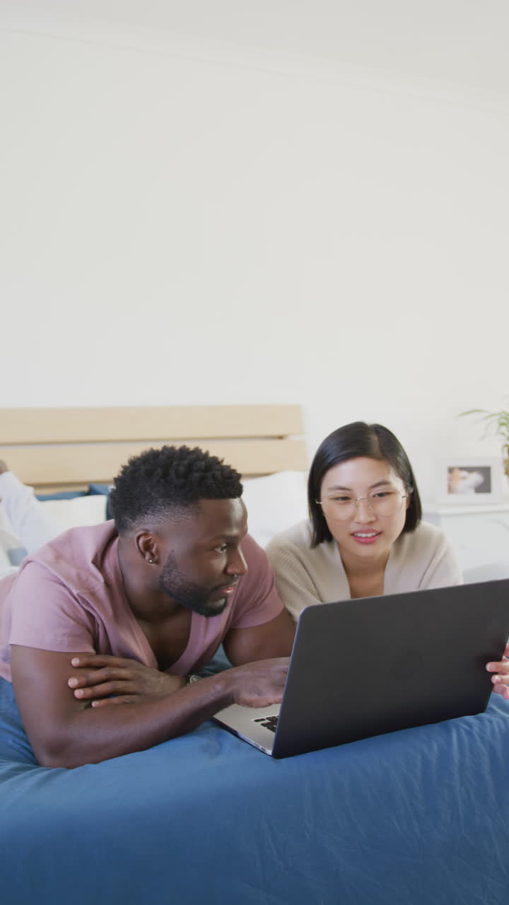 Vertical video of happy diverse couple lying and using laptop in bedroom