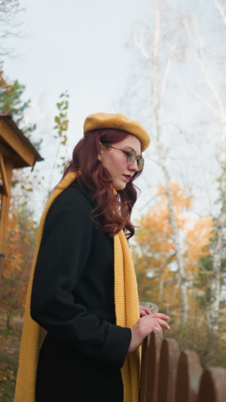 боковой вид леди в желтом берете, идущей к декоративному деревянному забору, отдыхая на руке, глядя на что-то на расстоянии с беседкой, осенними деревьями, сухими листьями и рекой на заднем плане