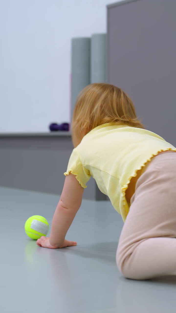 малыш держит теннисные мячи, один отскакивает от руки, в то время как взрослый опирается на зеленый мяч для упражнений в тренажерном зале, фитнес-среда включает в себя фиолетовый коврик, тренировочное оборудование и современный декор