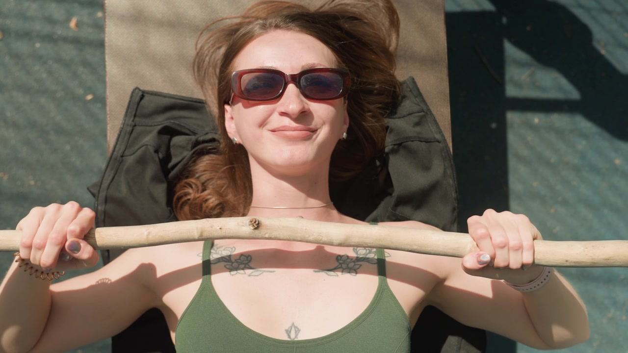 Close up of woman lying on her back outdoors holding onto wooden stick with both hands. Woman wears sunglasses and has visible tattoos across chest while resting on outdoor surface