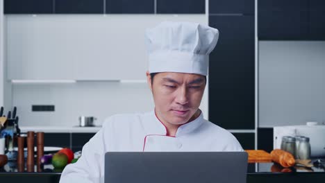 close up de un chef asiático usando una computadora portátil mientras está de pie en la cocina de casa